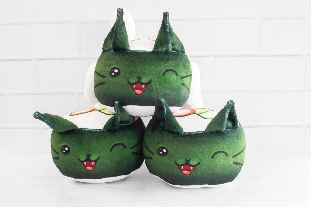 A handmade green plushie cat in the shape of a sushi roll with green ears and a white tail sticking out. It has a kawaii face with one eye winking and an open pink mouth that shows cat fangs. The top and the bottom have a white base and an image of a california roll's fillings in the shape of a heart. 