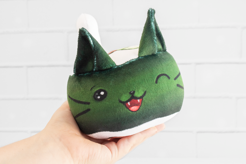 A handmade green plushie cat in the shape of a sushi roll with green ears and a white tail sticking out. It has a kawaii face with one eye winking and an open pink mouth that shows cat fangs. The top and the bottom have a white base and an image of a california roll's fillings in the shape of a heart. 