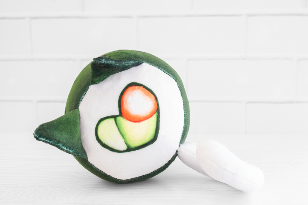 A handmade green plushie cat in the shape of a sushi roll with green ears and a white tail sticking out. It has a kawaii face with one eye winking and an open pink mouth that shows cat fangs. The top and the bottom have a white base and an image of a california roll's fillings in the shape of a heart. 