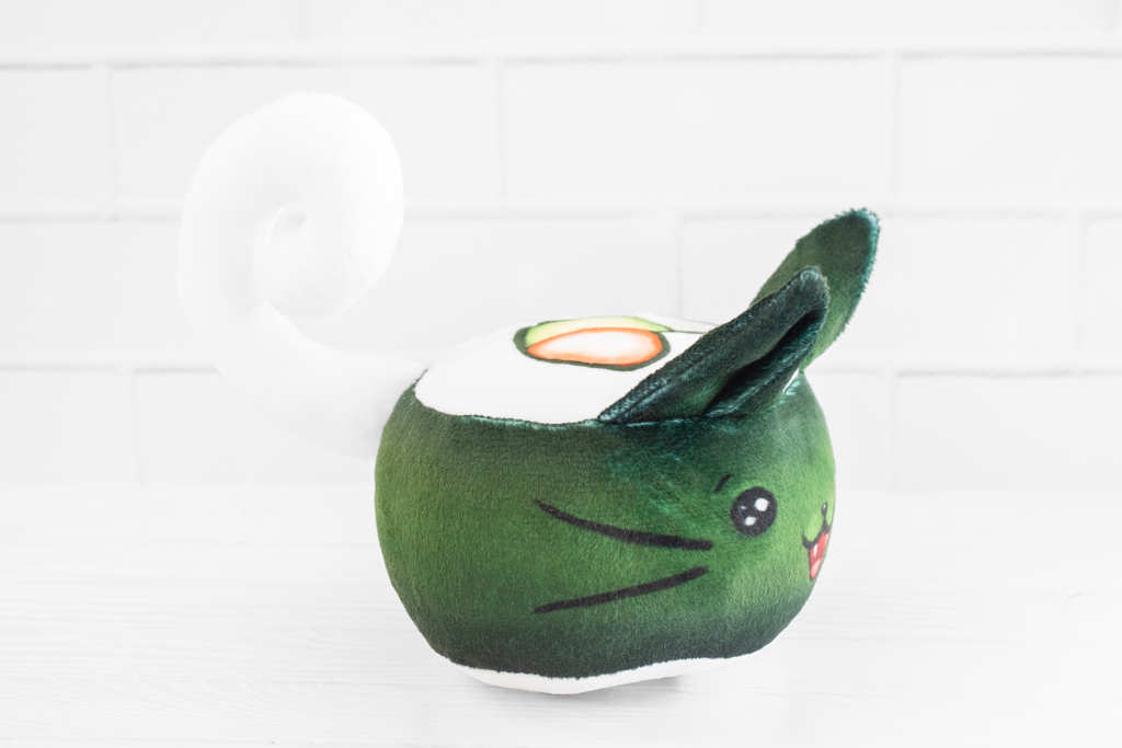 A handmade green plushie cat in the shape of a sushi roll with green ears and a white tail sticking out. It has a kawaii face with one eye winking and an open pink mouth that shows cat fangs. The top and the bottom have a white base and an image of a california roll's fillings in the shape of a heart. 