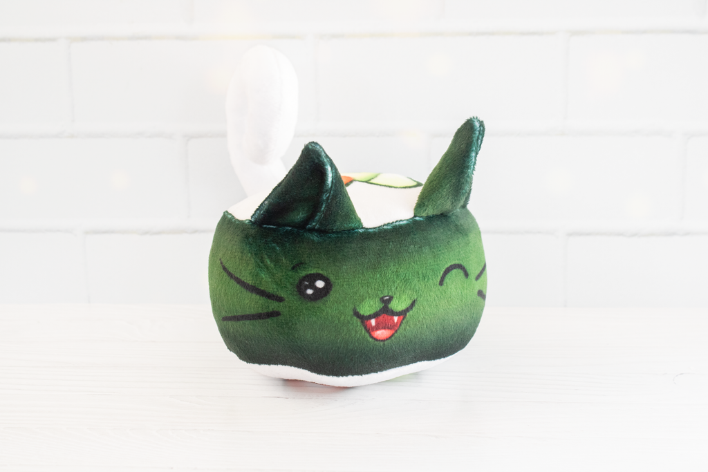 A handmade green plushie cat in the shape of a sushi roll with green ears and a white tail sticking out. It has a kawaii face with one eye winking and an open pink mouth that shows cat fangs. The top and the bottom have a white base and an image of a california roll's fillings in the shape of a heart. 