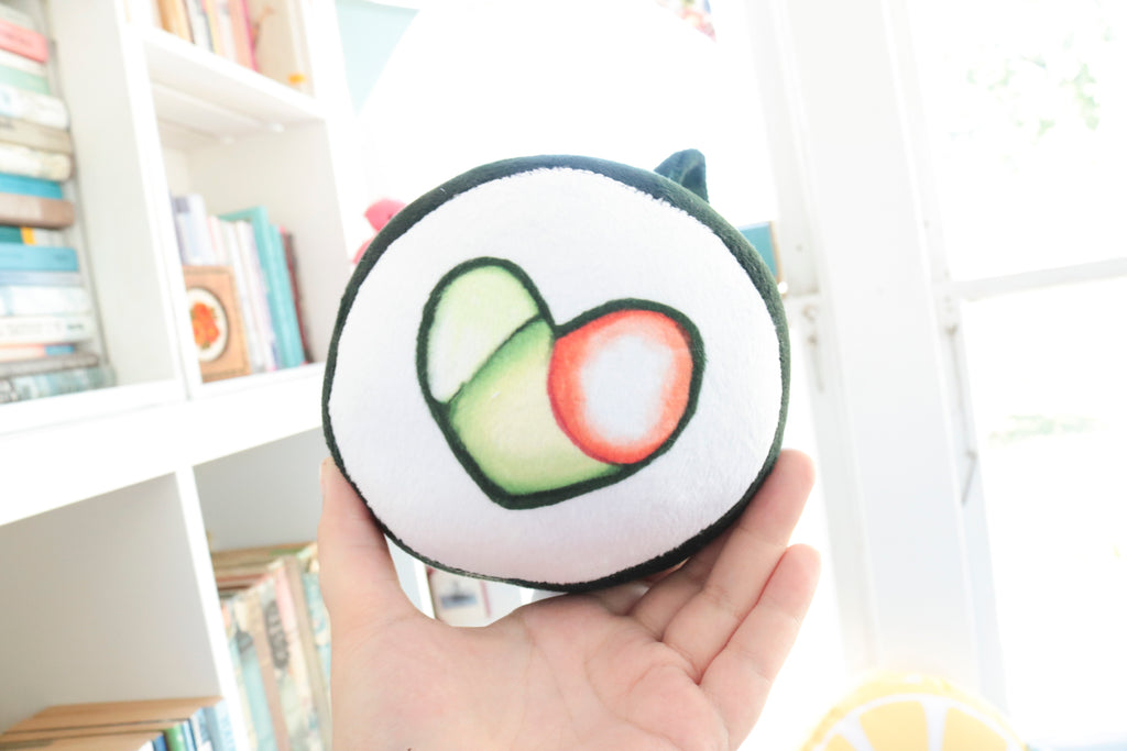 A handmade green plushie cat in the shape of a sushi roll with green ears and a white tail sticking out. It has a kawaii face with one eye winking and an open pink mouth that shows cat fangs. The top and the bottom have a white base and an image of a california roll's fillings in the shape of a heart. 