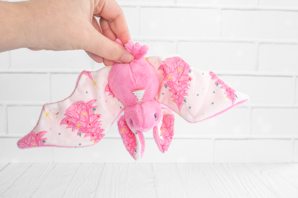 Handmade pink bat plush with vintage christmas tree print on the wings and white christmas tree charm at the neck. The eyes are pink and sparkly with snowflakes.