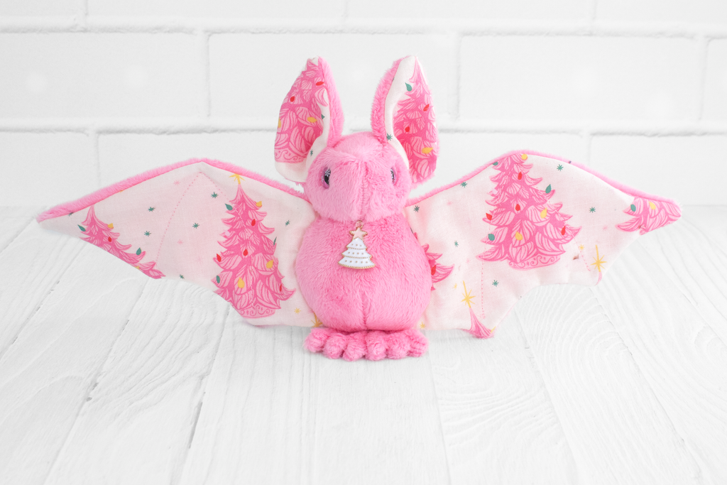 Handmade pink bat plush with vintage christmas tree print on the wings and white christmas tree charm at the neck. The eyes are pink and sparkly with snowflakes.
