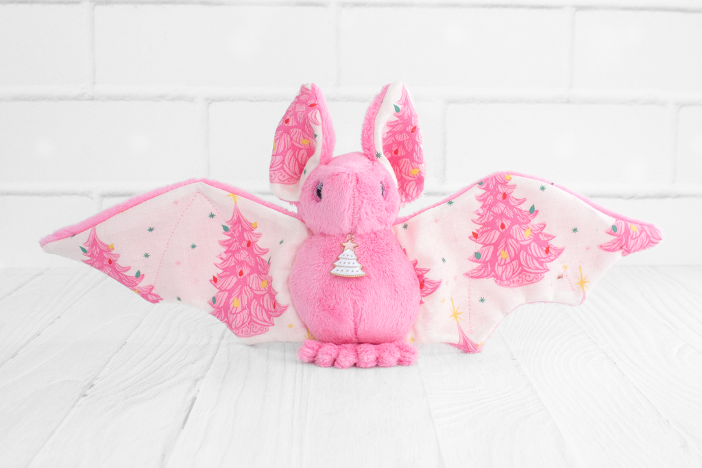 Handmade pink bat plush with vintage christmas tree print on the wings and white christmas tree charm at the neck. The eyes are pink and sparkly with snowflakes.