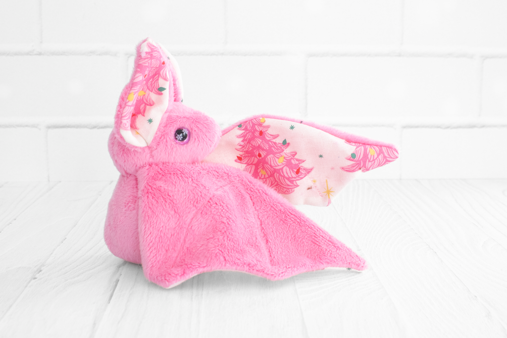 Handmade pink bat plush with vintage christmas tree print on the wings and white christmas tree charm at the neck. The eyes are pink and sparkly with snowflakes.