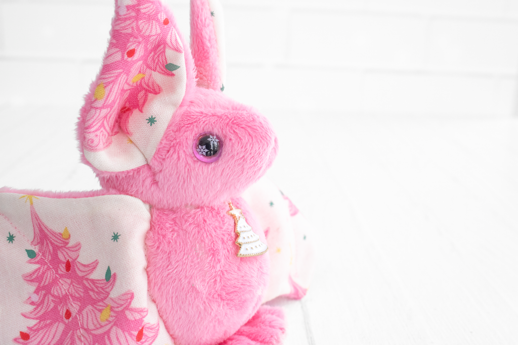 Handmade pink bat plush with vintage christmas tree print on the wings and white christmas tree charm at the neck. The eyes are pink and sparkly with snowflakes.