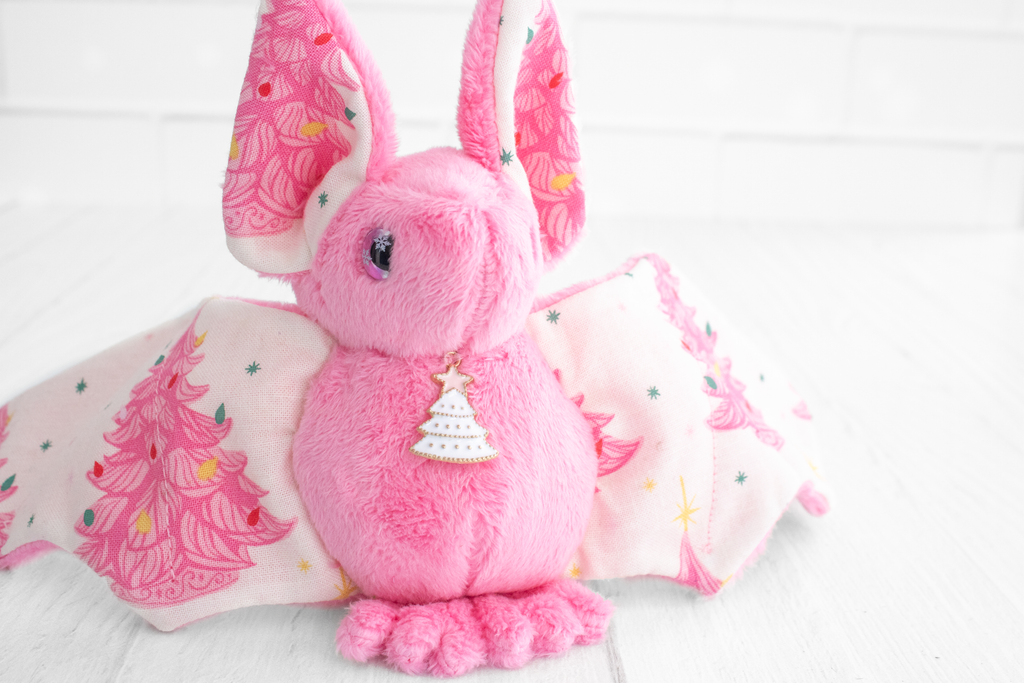 Handmade pink bat plush with vintage christmas tree print on the wings and white christmas tree charm at the neck. The eyes are pink and sparkly with snowflakes.