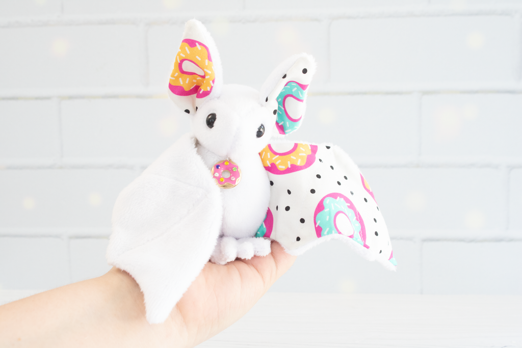 White plush bat with wings and ears patterned in colorful donuts and black polka dots, featuring a small matching donut charm on its chest, sitting against a light background.