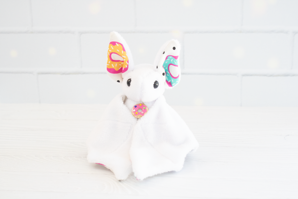 White plush bat with wings and ears patterned in colorful donuts and black polka dots, featuring a small matching donut charm on its chest, sitting against a light background.