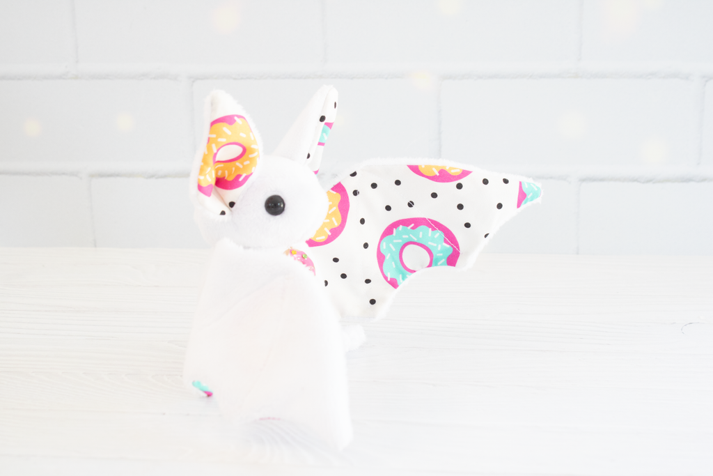 White plush bat with wings and ears patterned in colorful donuts and black polka dots, featuring a small matching donut charm on its chest, sitting against a light background.